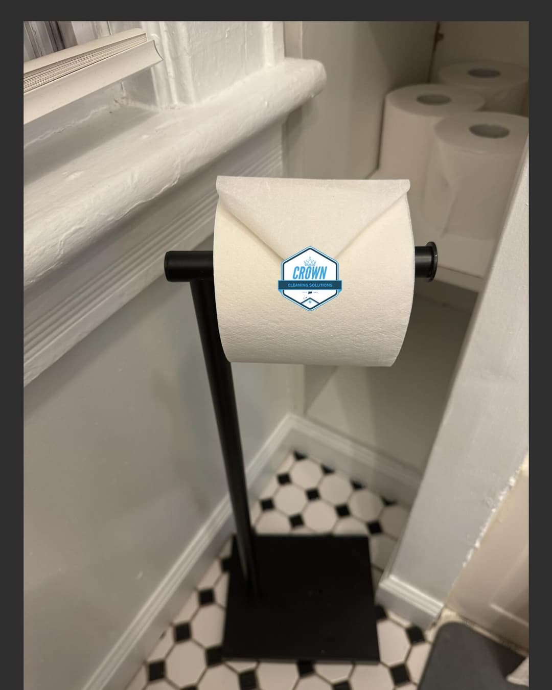 Toilet paper roll on a black stand in a bathroom with patterned tile floor. Crown logo visible.